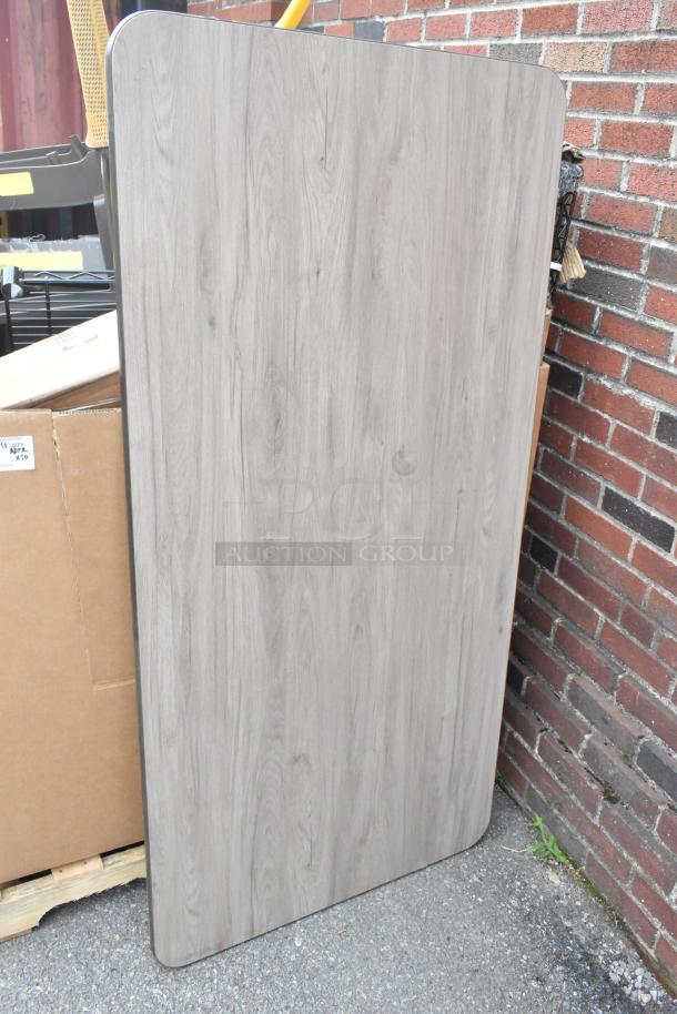 Brand new gray tabletop with wood pattern, scratch and dent condition, rectangular shape, displayed leaning on a wall.