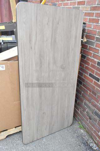 Brand new gray tabletop with wood pattern, scratch and dent condition, rectangular shape, displayed leaning on a wall.