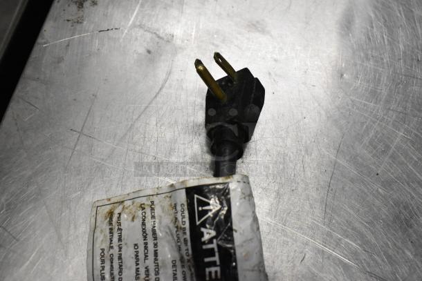 Close-up of a power plug against a stainless steel surface, with a partially visible warning label attached.