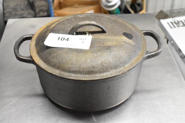 Cast iron pot with lid, used condition, visible rust on surface, numbered tag "104" attached to handle.