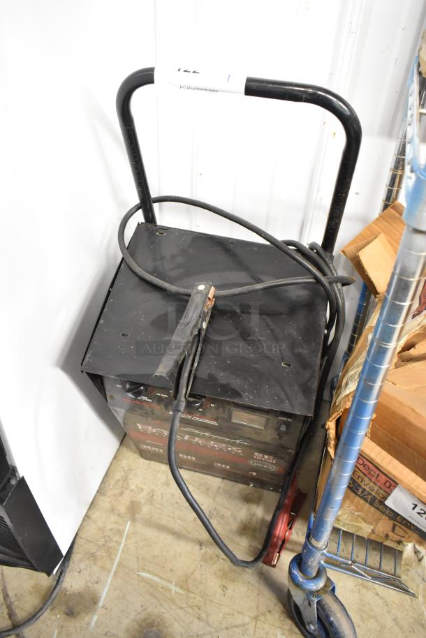Schumacher SE 8050 Battery Charger with attached cables, in used condition, black metal casing, on wheels.