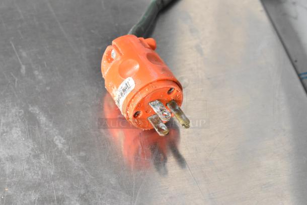 Orange power plug with visible metal prongs, laying on a metallic surface. Slight wear and label sticker present.