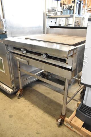 Stainless steel commercial gas-powered flat top griddle on casters, showing signs of wear, adjustable knobs, sturdy frame.