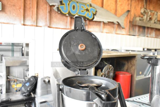 Stainless steel air pot with open lid, showing pump mechanism, in used condition, located in an industrial setting.