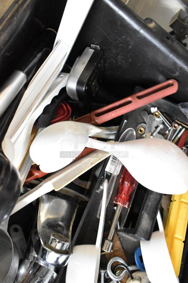 Assorted utensils and tools in a black poly bus bin, including white spoons, a screwdriver, and various small items.