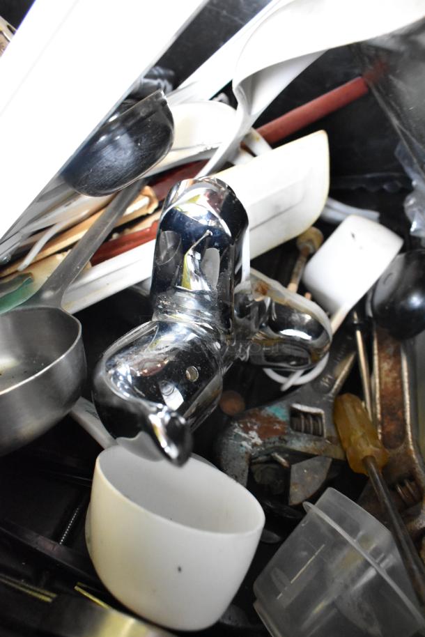 Black poly bus bin containing assorted utensils and tools, including chrome fittings, measuring cups, and screwdrivers. Condition varies.