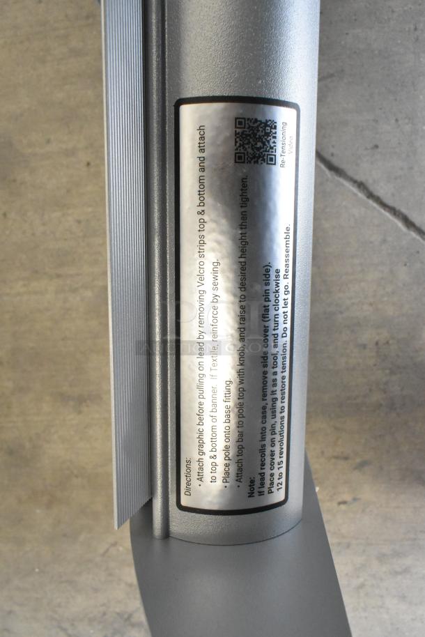 Roll-up sign base with attached instructions label showing setup directions and a QR code for retensioning video.
