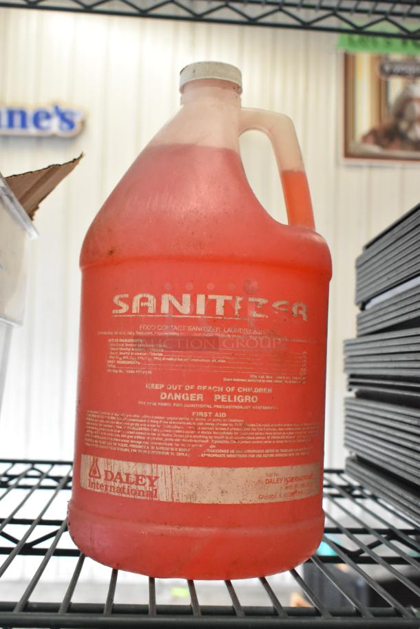 A gallon container of red sanitizer labeled "Daley International" on a metal shelf, part of a mixed lot of items.