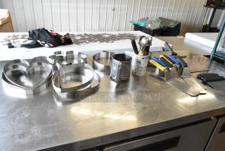 Lot of kitchen tools: spatulas, cake collars, heart-shaped cutters, paddle, and measuring cup on a stainless steel surface.