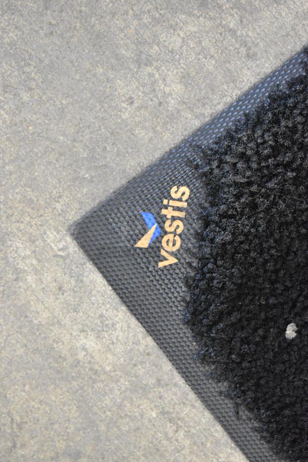 Black floor mat with textured surface and "vestis" logo on corner. Durable design, ideal for high-traffic areas.