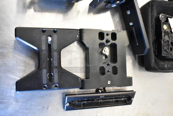 Four black metal brackets with adjustable slots and screws, partially placed on a black bag, showcasing industrial hardware components.