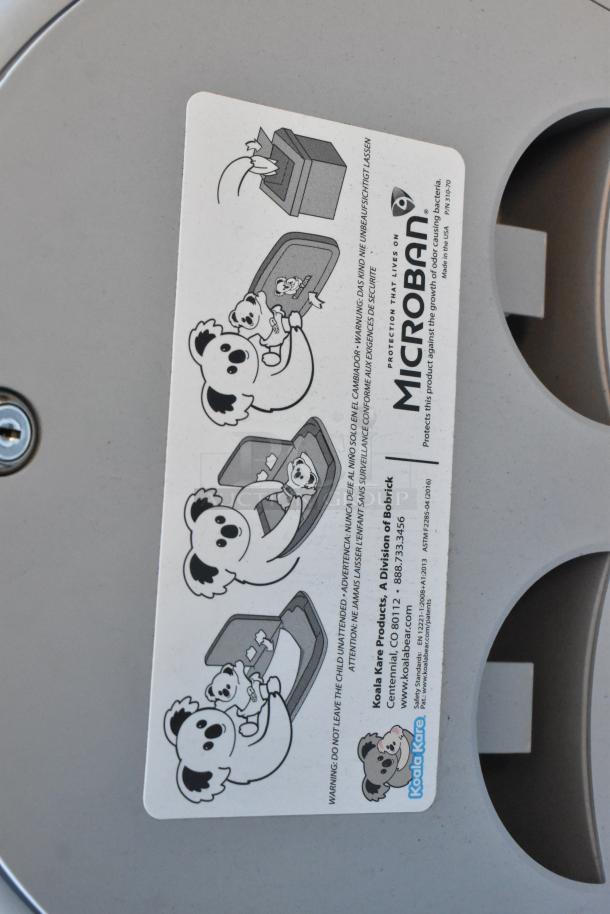 Koala Kare gray wall mount diaper changing station, durable plastic, safety instructions visible, featuring Microban protection.