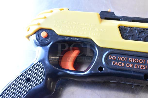 Bugasalt gun close-up with safety label and patent info. Features yellow/black design, visible trigger, and caution text.