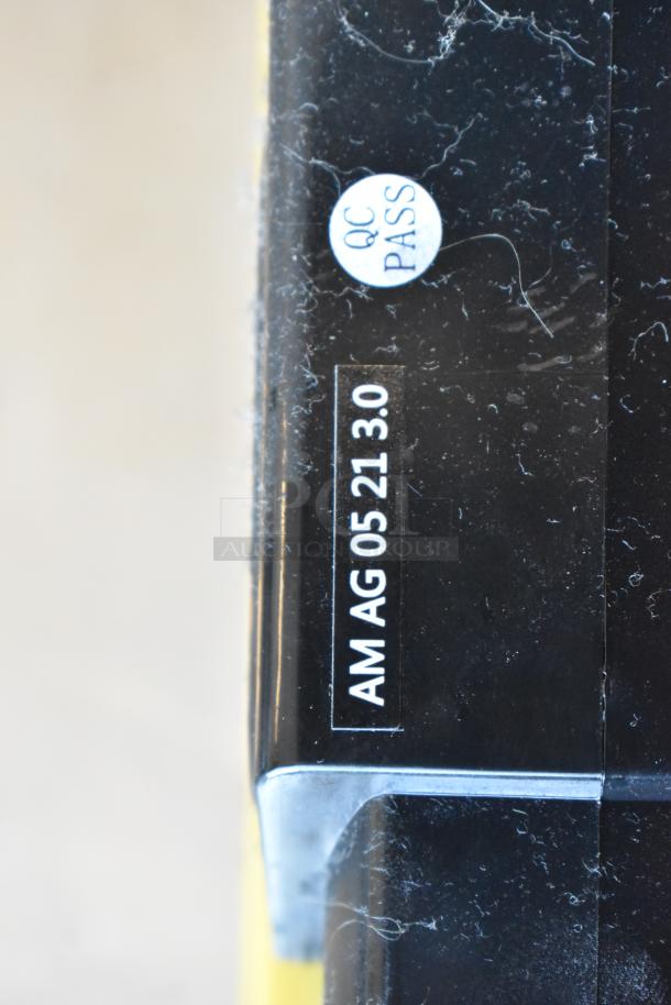 Close-up of a black item's side with a "QC Pass" sticker and marking "AM AG 05 21 3.0," indicating quality control approval.
