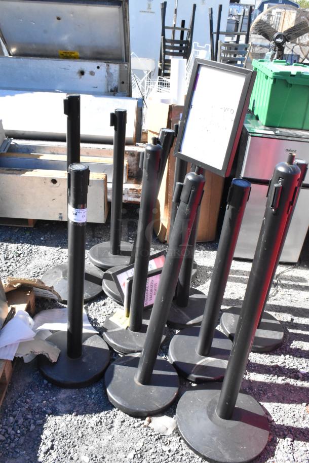 10 QueueWay stanchions in used condition, black finish, solid bases, various labels attached; ideal for crowd control.