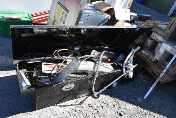 Black diamond plate pickup truck box with contents, including tools and metal items. Shows open lid and various fittings.