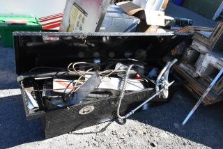 Black diamond plate pickup truck box with contents, including tools and metal items. Shows open lid and various fittings.