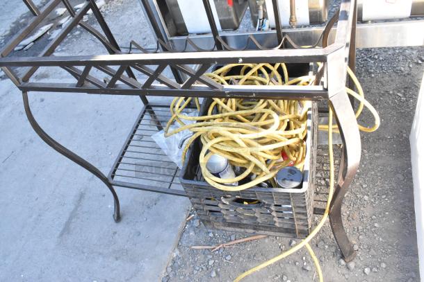 Metal frame with decorative design, holds a plastic crate containing yellow extension cords and spray cans, on outdoor gravel surface.