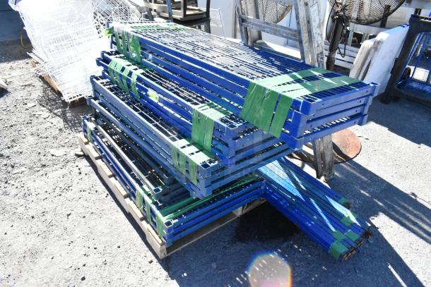 Lot of 18 blue finish metal wire shelves with poles, stacked outdoors on pallets, secured with green straps.