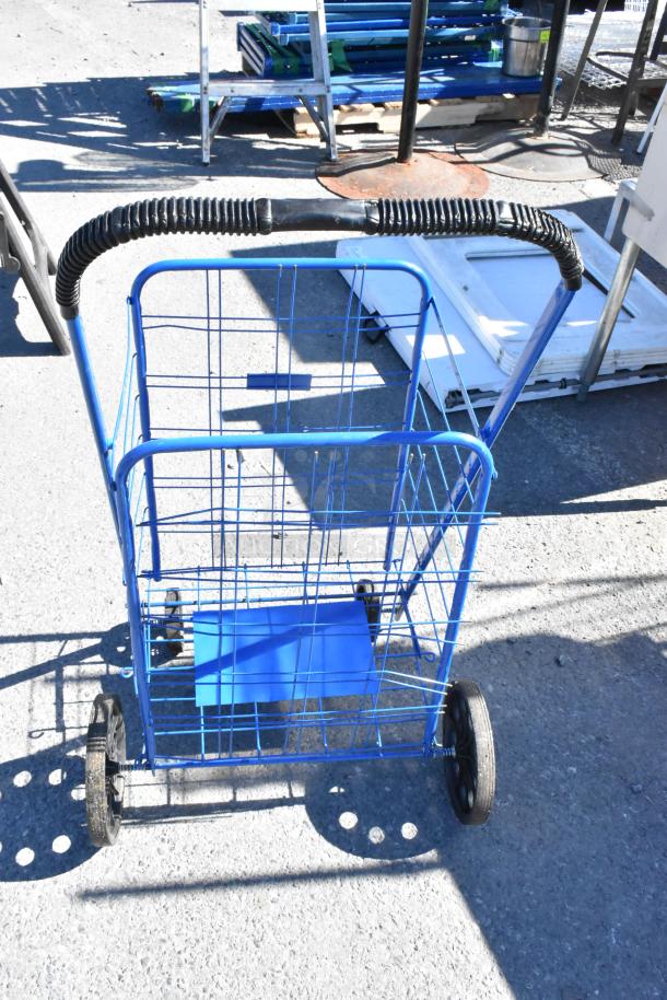 Blue wire basket cart with black handle and wheels, shown outdoors on paved surface. Used condition with visible wear.