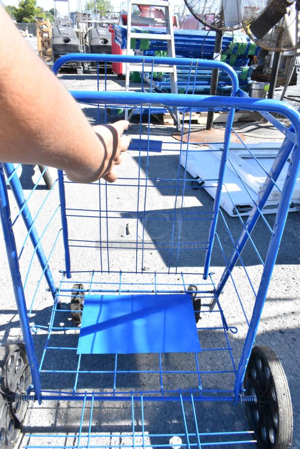Blue metal cart with black wheels, visible in an outdoor setting. Surrounded by industrial items and concrete surface.