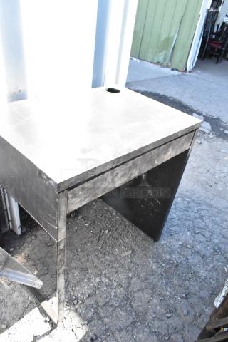 Black metal desk with a worn surface, featuring a cable management hole on the top, set on concrete outdoors.