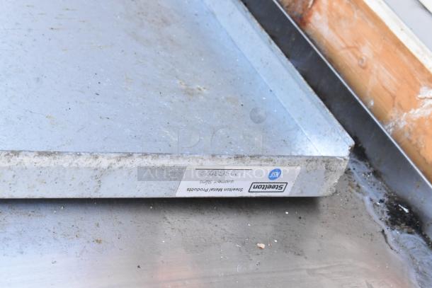 Stainless steel table with visible wear and signs of use. Label reads "Steelfon Metal Products, Equipment Stand."