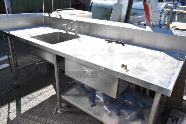 Stainless steel table with sink basin, dual-handle faucet, drawer, under shelf, and protective backsplash.