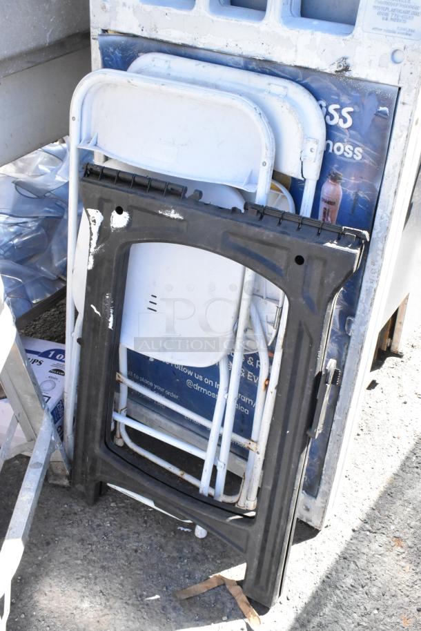 Ladder, folding chairs, saw horse, and table stacked outdoors, showing signs of wear.