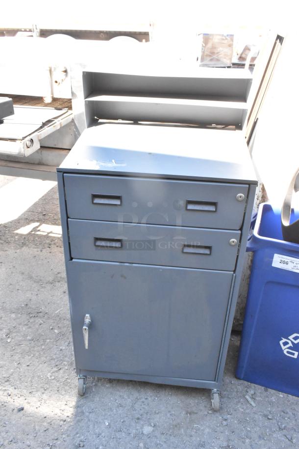Gray metal cabinet with 2 drawers on commercial casters, featuring a locking mechanism and shelf, in used condition.