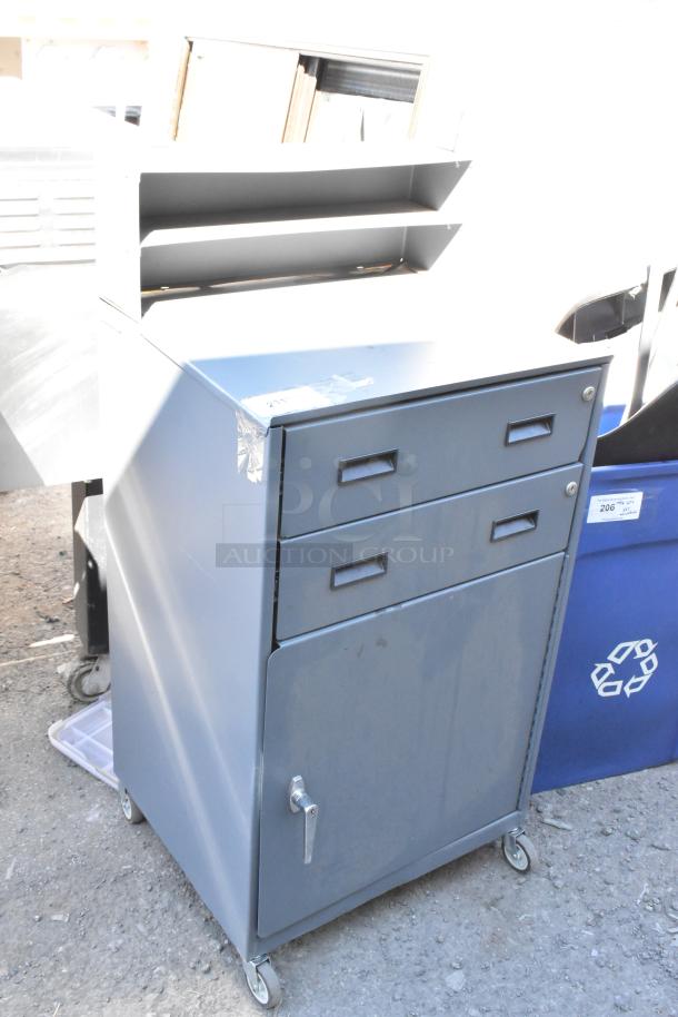 Gray metal cabinet with two drawers and door, on commercial casters. Shows slight wear, includes shelving unit on top.