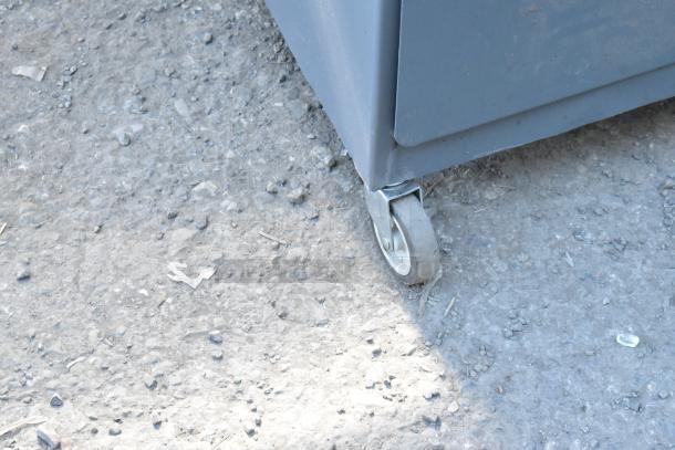 Gray metal cabinet corner with commercial caster wheel on textured ground. Appears in functional condition.