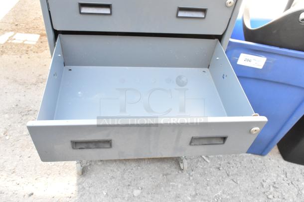 Gray metal cabinet with two drawers, shown on commercial casters. Visible wear. Empty opened drawer, suitable for storage.