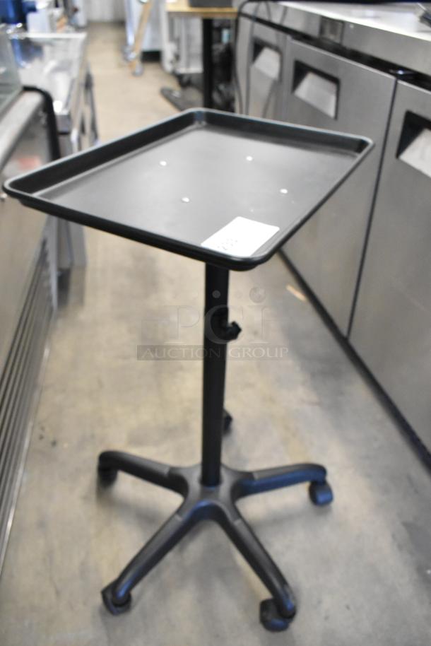 Black tray dolly on casters, adjustable height, in good condition. Features a square tray top and five-wheel base.