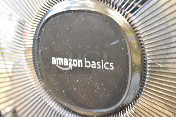 Amazon Basics floor fan, 120V, 1 phase, close-up view of the logo and fan blades. Dust visible on surface.