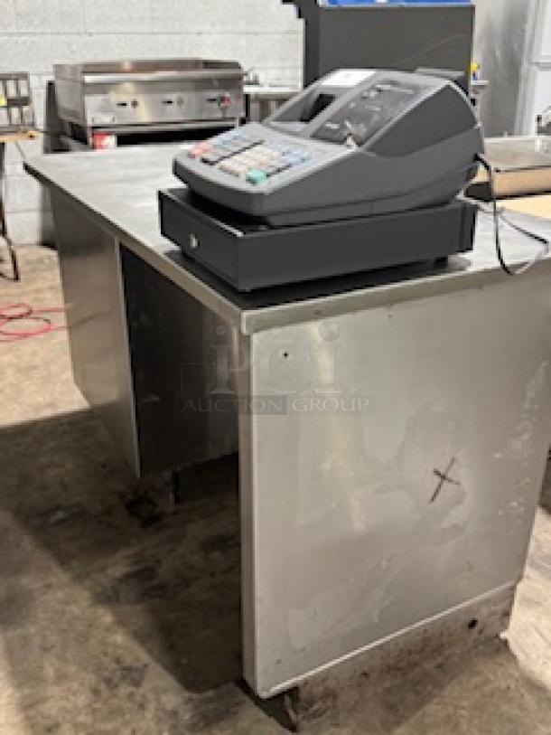 Stainless steel counter by S&G Manufacturing, Model BC7-1543, with attached cash register. Visible wear, sturdy condition.