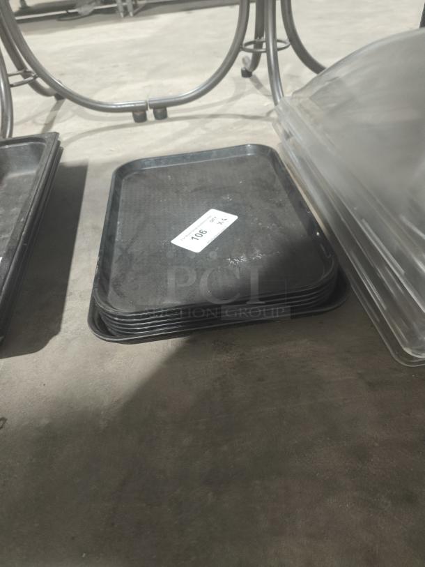 Stack of black commercial plastic cafeteria trays, each 18" x 14", marked with auction number 106. Trays appear in used condition.
