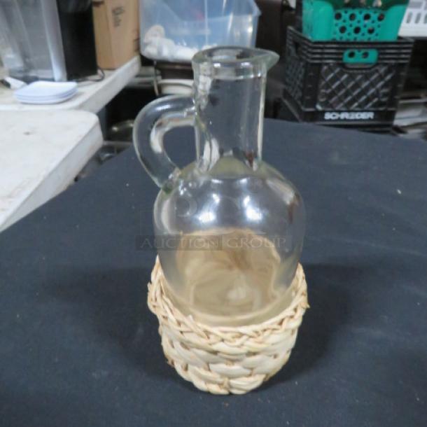 Clear glass vinegar bottle in a light straw basket holder, featuring a rounded body and a handle, in good condition.
