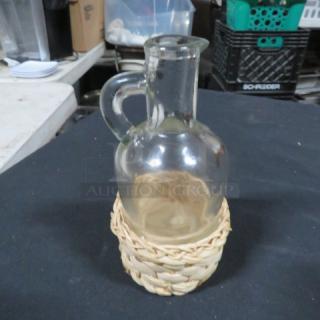 Clear glass vinegar bottle in a woven straw basket holder. Features a handle and narrow spout. Good condition.