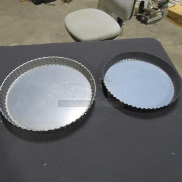Assorted bake pans, two round fluted-edge pans on dark surface; one silver, one black; minimal wear visible.