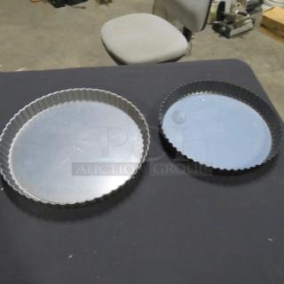 Assorted bake pans, two round fluted-edge pans on dark surface; one silver, one black; minimal wear visible.