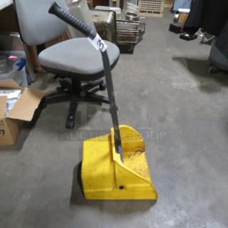 Yellow commercial dust pan with long black handle, moderate wear, visible scuff marks. Ideal for industrial cleaning tasks.