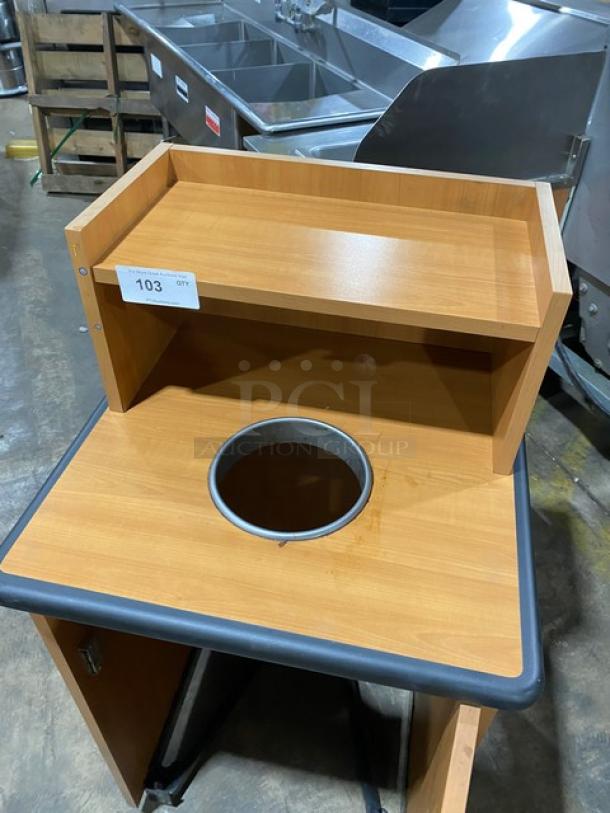 Commercial wooden recycle-style receptacle cabinet with upper tray storage, on legs. Features waste disposal opening and storage shelf.