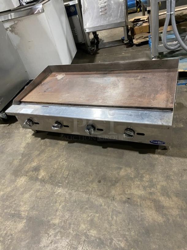 Cook Rite stainless steel natural gas countertop flat griddle, with back and side splash, on legs. Four control knobs.