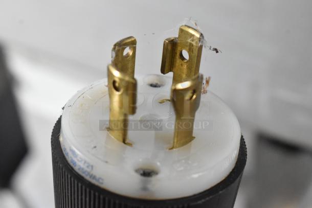 Close-up of a plug or connector, showing metal prongs and a partially visible inscription indicating voltage details.