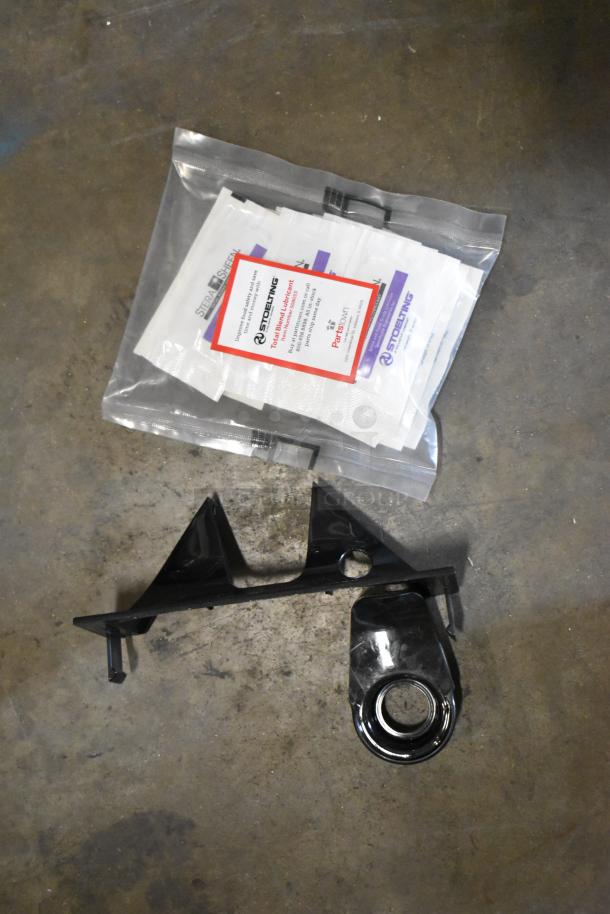 Packaged parts for Stoelting SCBF128-37 machine, including lubricant pack and black plastic bracket on a concrete surface.