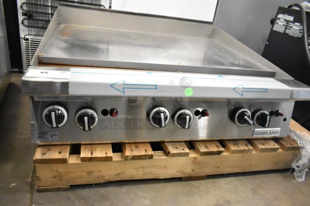 BRAND NEW Garland Stainless Steel Commercial Flat Top Griddle, minor scratches, four control knobs, on wooden pallet.
