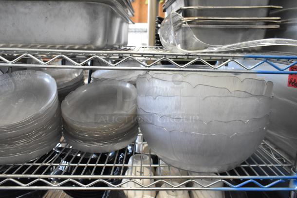 Assorted kitchen items including stainless steel drop-in bins, chafer lids, salad plates, and bowls on metal shelving.
