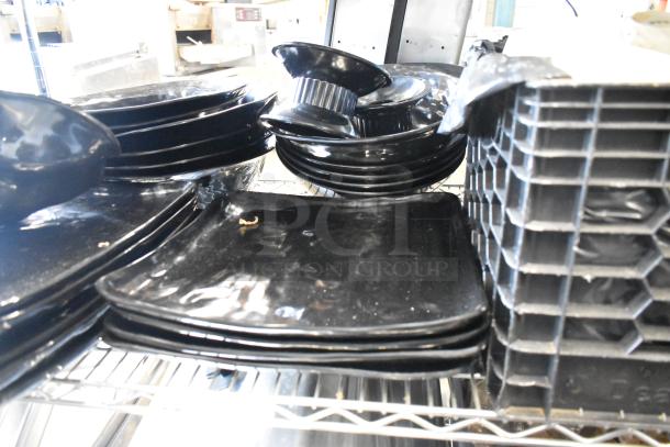 Metro lot of items including stainless steel drop-in bins, chafer lids, black salad plates, and bowls on a shelf. No shelving unit.