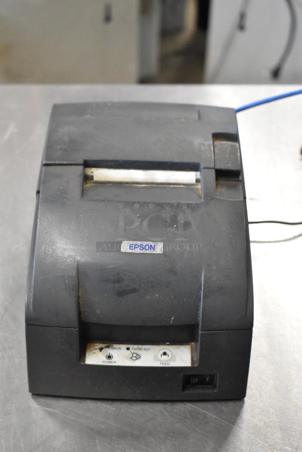 Epson receipt printer, used, black, front view showing feed button and power indicator, slight wear visible.